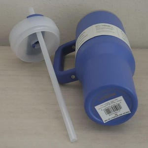 Puede incluir: Un vaso t&eacute;rmico azul con asa y tapa blanca con pajita. El vaso tiene una etiqueta blanca enrollada. La pajita es blanca e inclinada hacia la tapa. El vaso est&aacute; sobre una superficie clara.