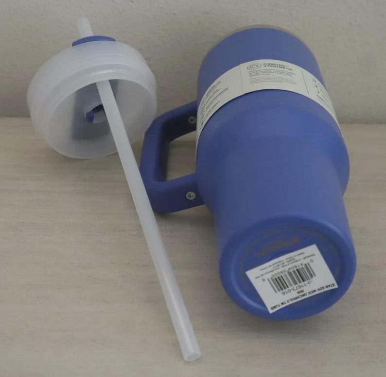 Puede incluir: Un vaso t&eacute;rmico azul con asa y tapa blanca con pajita. El vaso tiene una etiqueta blanca enrollada. La pajita es blanca e inclinada hacia la tapa. El vaso est&aacute; sobre una superficie clara.