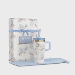 May include: A white Stanley travel mug and gift box set. The mug and box are decorated with a floral and bow pattern in pink and blue. The mug has a blue handle and lid, and the box is tied with a blue ribbon.