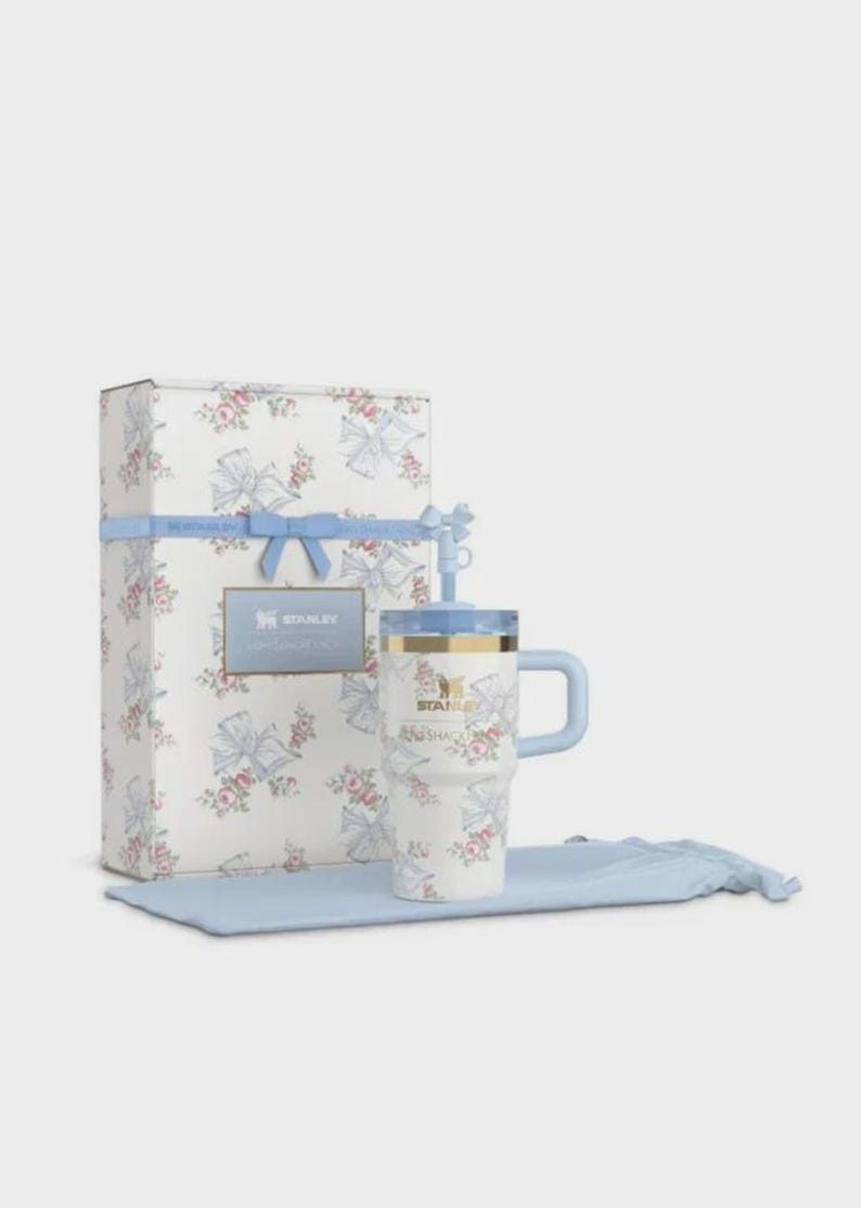 May include: A white Stanley travel mug and gift box set. The mug and box are decorated with a floral and bow pattern in pink and blue. The mug has a blue handle and lid, and the box is tied with a blue ribbon.