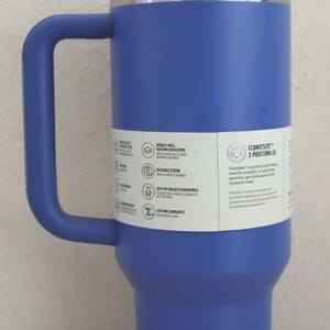 Puede incluir: Un vaso t&eacute;rmico azul con tapa y asa blancas. El vaso tiene una pajita blanca y un borde de acero inoxidable. Una etiqueta en el lateral enumera caracter&iacute;sticas como aislamiento al vac&iacute;o de doble pared y pajita reutilizable. La tapa est&aacute; etiquetada como "FlowState 3-Position Lid."