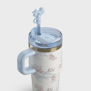 May include: A white floral tumbler featuring pink roses and grey bows. It has a light blue handle, lid, and straw. The tumbler has a gold band near the top.
