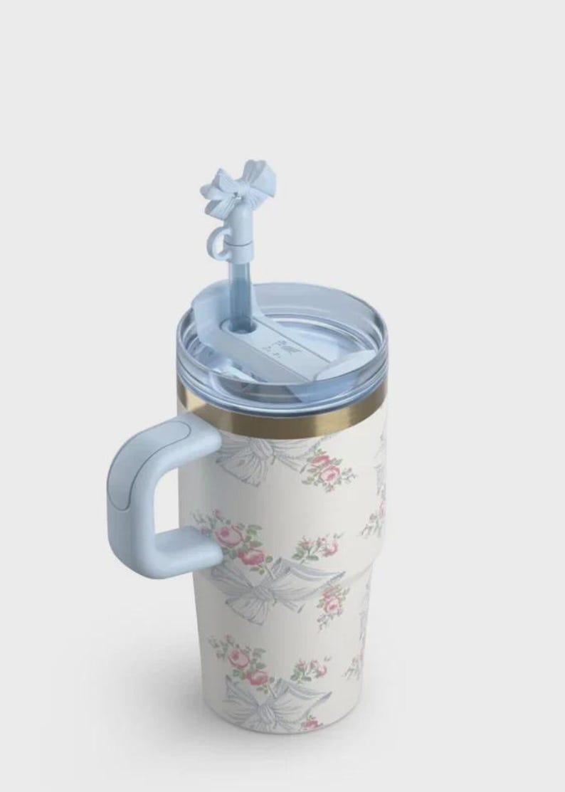 May include: A white floral tumbler featuring pink roses and grey bows. It has a light blue handle, lid, and straw. The tumbler has a gold band near the top.