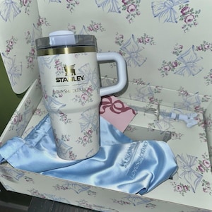 May include: A white Stanley travel mug with a floral and bow pattern, a white handle, and a clear lid. The mug is inside a box with a matching floral design, with a blue satin bag and a pink tag.