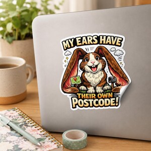 May include: A die-cut sticker featuring a brown and white rabbit with long ears, a small green map, and a mailbox. The text reads MY EARS HAVE THEIR OWN POSTCODE! in bold yellow and black letters against a white background on a laptop surface.