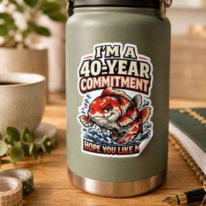May include: A matte olive green stainless steel water bottle featuring a sticker with a red, white, and black koi fish illustration. The text reads I'M A 40-YEAR COMMITMENT HOPE YOU LIKE ME in bold, stylized lettering above and below the fish.