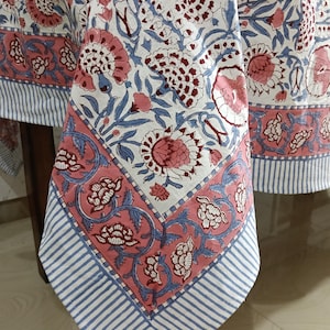 Könnte beinhalten: Eine weiße Tischdecke mit einem floralen Muster in Rosa und Kastanienbraun, akzentuiert mit blauen Blättern und Ranken. Der Rand hat ein rosa Band mit floralen Designs und blau-weißen Streifen. Die Tischdecke liegt auf einem Holztisch.