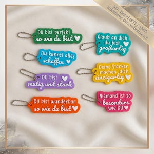 Könnte beinhalten: Eine Auswahl an farbenfrohen Schlüsselanhängern mit positiven Affirmationen auf Deutsch. Jeder Schlüsselanhänger hat eine andere Farbe und Form, mit Sätzen wie "Du bist perfekt, so wie du bist" und "Glaube an dich, du bist großartig."