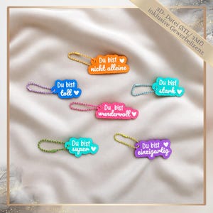 Puede incluir: Una colección de llaveros coloridos con frases alemanas y símbolos de corazón. Los llaveros son de varias formas y colores, incluyendo naranja, azul, rosa y morado. Cada llavero tiene una cadena de metal.