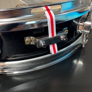 May include: Close-up of a black snare drum with chrome hardware. A white ribbon with red stripes is attached to the drum. The drumhead is a light beige colour.