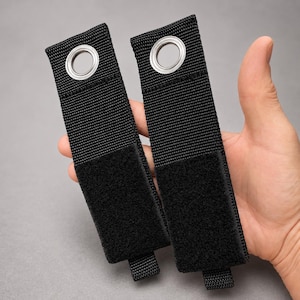 May include: Two black nylon straps with metal grommets and hook-and-loop fasteners. The straps are held in a hand, showcasing their design and construction. The grommets are silver, and the hook-and-loop fasteners are black.