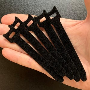May include: A close-up shot of several black hook and loop cable ties. Each tie has a rectangular body with a rounded end and a small hole. The ties are arranged in a fan shape, and the image is well-lit.