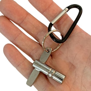 May include: A silver drum key attached to a key ring and a black carabiner clip. The drum key has a T-shaped handle and a cylindrical body. The carabiner clip is black with a silver accent. The key ring is gold-colored.