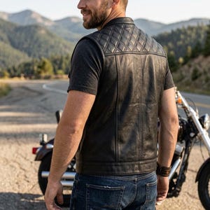 May include: A black leather vest featuring a quilted diamond-patterned yoke on the upper back and shoulders. The vest has a smooth leather lower back panel, a stand-up collar, and vertical seam detailing, worn over a dark gray short-sleeve t-shirt.