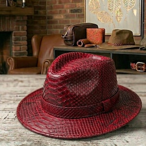 Può includere: Un cappello fedora rosso con un motivo a pelle di serpente. Il cappello ha una tesa larga e una corona strutturata. Il cappello è realizzato in un materiale strutturato ed è di una tonalità di rosso intenso. È un accessorio elegante.