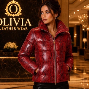Può includere: Un piumino rosso con motivo a pelle di serpente nera, collo alto e cerniera frontale a tutta lunghezza. La giacca presenta una consistenza trapuntata e maniche lunghe. Il testo OLIVIA LEATHER WEAR è visibile sullo sfondo dietro la giacca.