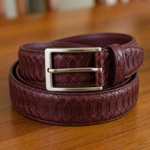 Puede incluir: Cinturón de cuero burdeos con un patrón de piel de serpiente texturizado. Tiene una hebilla rectangular plateada y está enrollado sobre una superficie de madera. Un accesorio clásico.