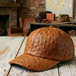 May include: A brown leather baseball cap with a textured surface. The cap has a curved brim and a rounded crown. The cap is made of a material that appears to be ostrich leather. The cap is sitting on a wooden surface.