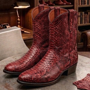 May include: A pair of deep red cowboy boots featuring a textured snakeskin pattern across the entire exterior. The boots have a pointed toe, a mid-calf height, pull tabs at the top, and a stacked heel. The interior lining is a light tan color.