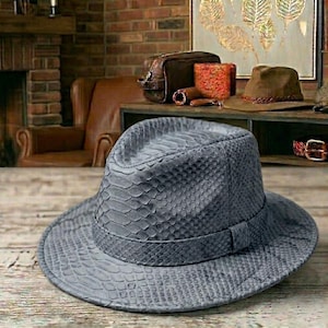 Könnte beinhalten: Ein grauer Fedora-Hut mit Schlangenmuster. Der Hut hat eine strukturierte Krone und eine breite Krempe. Der Hut besteht aus einem strukturierten Material und liegt auf einer Holzoberfläche. Andere Hüte und Accessoires befinden sich im Hintergrund.