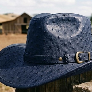 Pode incluir: Um chapéu de cowboy azul marinho com uma superfície texturizada e uma faixa de couro. O chapéu apresenta uma fivela dourada e está situado contra um cenário rural com uma cerca de madeira e um celeiro.
