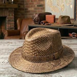 Può includere: Un cappello fedora marrone con motivo a pelle di serpente. Il cappello ha un'ampia tesa e una fascia intorno alla corona. Altri accessori in pelle sono visibili sullo sfondo, tra cui un portafoglio, un beauty case e una cintura.