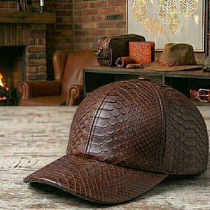 Puede incluir: Gorra de béisbol de cuero marrón con un patrón texturizado similar a la piel de reptil. La gorra tiene una visera curva y una corona estructurada. Se muestra sobre una superficie de madera, con un fondo borroso de chimenea y otros artículos de cuero.