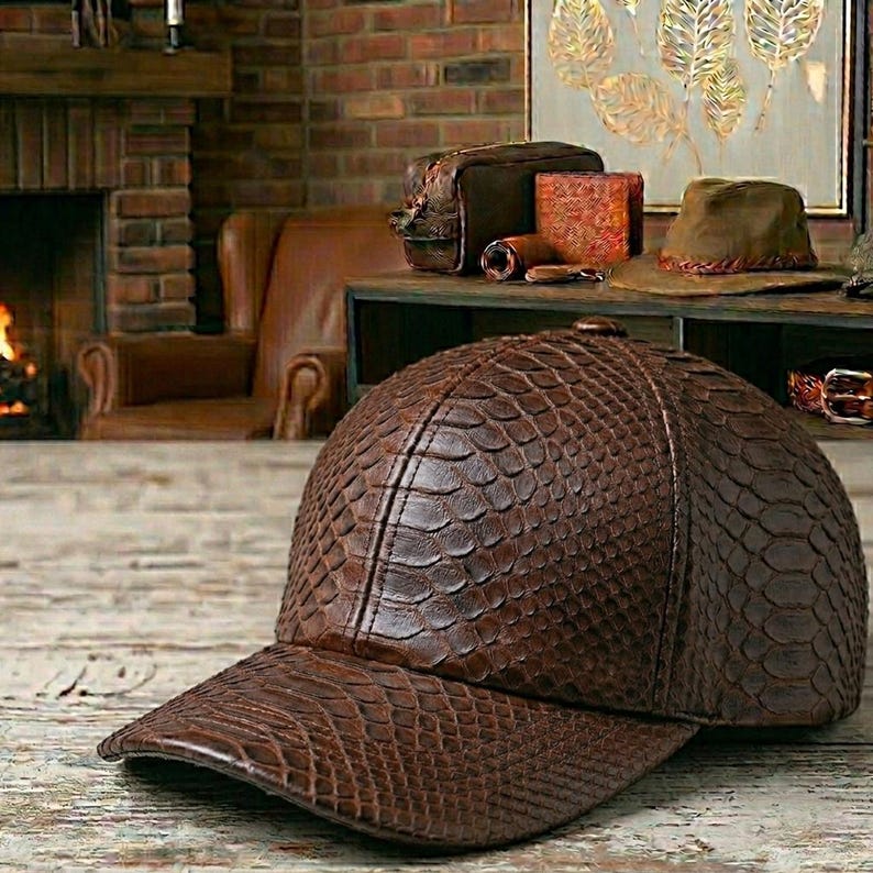 May include: A brown leather baseball cap with a reptile skin texture. The cap features a curved peak and a structured crown. The hat is displayed on a wooden surface, with a blurred background of a fireplace and other leather items.