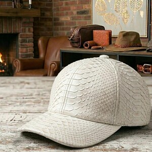 May include: A cream-colored baseball cap with a textured, scale-like pattern. The cap has a curved brim and a button on top. Other leather accessories, including a toiletry bag, wallet, and belt, are visible in the background.
