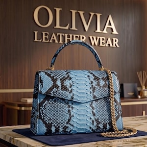 Peut inclure: Un sac à main structuré avec un motif peau de serpent bleu clair et noir, une poignée supérieure et une bandoulière en chaîne dorée. Le sac possède un rabat et est placé devant une enseigne murale indiquant OLIVIA LEATHER WEAR.