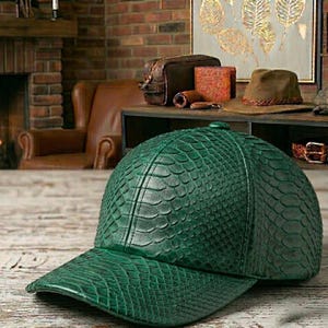 Puede incluir: Gorra de béisbol verde con un patrón texturizado similar a la piel de reptil. La gorra tiene una visera curva y un botón en la parte superior. Otros accesorios de cuero son visibles en el fondo.