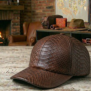 May include: A brown baseball cap with a textured, reptile-like pattern. The cap has a curved brim and a structured crown. The hat is made of a dark brown material and appears to be leather. Other leather goods are visible in the background.