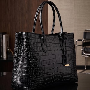 May include: A black tote bag with a glossy crocodile skin texture, featuring two upright handles and a small hanging luggage tag with a gold-toned metal accent at the base. The bag has a structured rectangular shape and a polished finish.