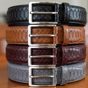 May include: A stack of four leather belts in black, brown, burgundy, and gray. Each belt features a textured snakeskin pattern and a silver-colored rectangular buckle. The belts are stacked vertically, showcasing their different colors and textures.