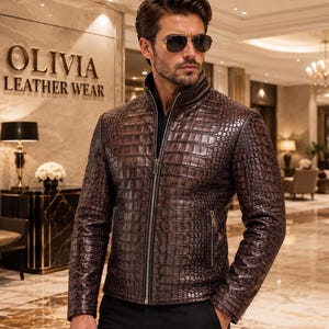 Puede incluir: Una chaqueta de cuero marrón oscuro con un patrón texturizado de piel de cocodrilo, que presenta un cuello alto, una cremallera frontal de metal plateado y dos bolsillos laterales con cremalleras, sobre una camisa negra y pantalones oscuros.