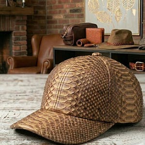 Puede incluir: Gorra de béisbol marrón con estampado de piel de serpiente. La gorra tiene una visera curva y un botón en la parte superior. Otros accesorios de cuero son visibles en el fondo, como un neceser, una cartera y un cinturón.