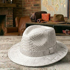 Peut inclure: Un chapeau fedora gris clair avec un motif peau de serpent. Le chapeau a une couronne structurée et un large bord. Le chapeau est fait d'un matériau texturé et comporte une bande assortie autour de la base de la couronne. D'autres accessoires en cuir sont en arrière-plan.