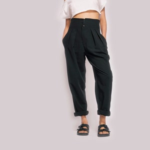 K&ouml;nnte beinhalten: Eine Frau tr&auml;gt ein wei&szlig;es, schulterfreies Crop-Top und eine schwarze, hochgeschlossene Hose mit Knopfverschluss. Sie tr&auml;gt schwarze Sandalen mit Schnallen.