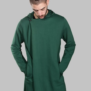Könnte beinhalten: Ein langärmliger, dunkelgrüner Kapuzenpullover mit einem einzigen Knopfverschluss am Halsausschnitt. Der Pullover hat eine lockere Passform und ein langes, fließendes Design.