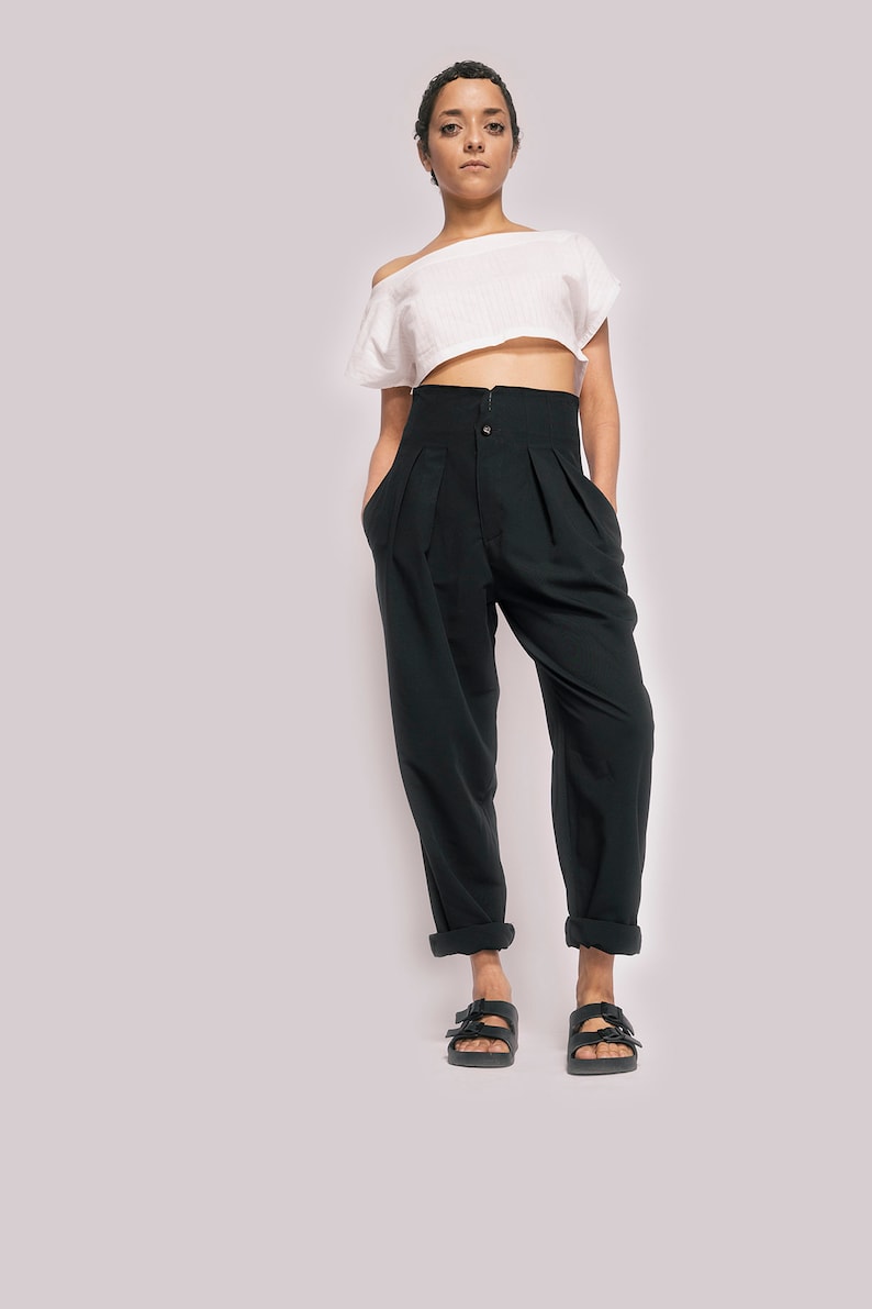 K&ouml;nnte beinhalten: Eine Frau tr&auml;gt ein wei&szlig;es, schulterfreies Crop-Top und eine schwarze, hochgeschlossene Hose mit Knopfverschluss. Sie tr&auml;gt schwarze Sandalen mit Schnallen.