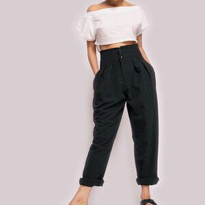 K&ouml;nnte beinhalten: Eine Frau tr&auml;gt ein wei&szlig;es, schulterfreies Crop-Top und eine schwarze, hochgeschlossene Hose mit Falten. Sie tr&auml;gt schwarze Sandalen.