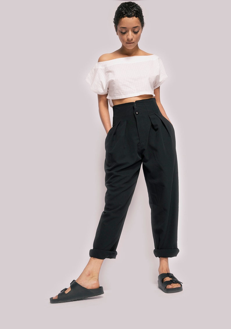 K&ouml;nnte beinhalten: Eine Frau tr&auml;gt ein wei&szlig;es, schulterfreies Crop-Top und eine schwarze, hochgeschlossene Hose mit Falten. Sie tr&auml;gt schwarze Sandalen.