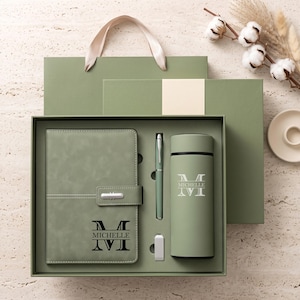 May include: A sage green gift set containing a notebook with a magnetic closure, a matching pen, a small USB drive, and a cylindrical insulated bottle. Each item is personalised with the letter M and the name Michelle in a dark font.