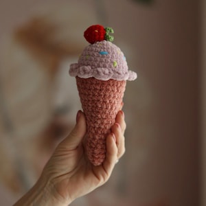Puede incluir: Un cono de helado de ganchillo con una fresa encima. El cono es de color melocotón claro, y el helado es de un lavanda suave con chispas de colores. La fresa es roja con hojas verdes.