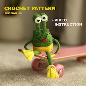 Könnte beinhalten: Eine handgefertigte grüne und gelbe Häkelfigur mit großen Augen und einem roten Lächeln, die auf einem rosa Skateboard sitzt. Das Bild enthält den Text "CROCHET PATTERN PDF ENGLISH" und "+ VIDEO INSTRUCTION".