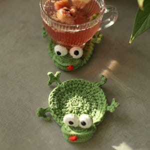 Puede incluir: Dos posavasos de rana verde de ganchillo con ojos blancos y lenguas rojas. Una taza de té de cristal transparente con una bebida roja se encuentra sobre uno de los posavasos. Los posavasos tienen cuatro patas pequeñas.