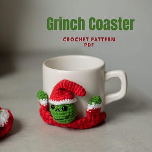 Puede incluir: Un posavasos de ganchillo con la cara de un personaje verde, un gorro de Papá Noel rojo y borde blanco, envuelto alrededor de una taza de cerámica blanca. El texto Grinch Coaster CROCHET PATTERN PDF aparece sobre la taza en letras verdes y rojas.