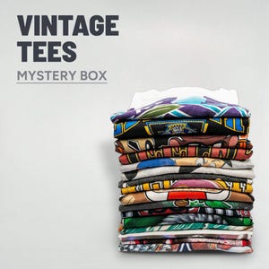 Op de afbeelding: Een stapel opgevouwen vintage t-shirts met diverse kleurrijke grafische ontwerpen. De shirts zijn netjes gerangschikt en tonen een verscheidenheid aan patronen en kleuren. Tekst bovenaan luidt "VINTAGE TEES" en "MYSTERY BOX".