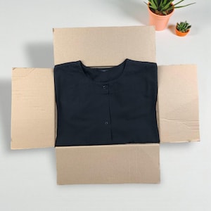 Może przedstawiać: Czarna koszula zapinana na guziki jest starannie złożona w otwartym kartonowym pudełku. Koszula ma okrągły dekolt i pojedynczy rząd guzików z przodu. W tle widoczne są dwie małe rośliny doniczkowe.