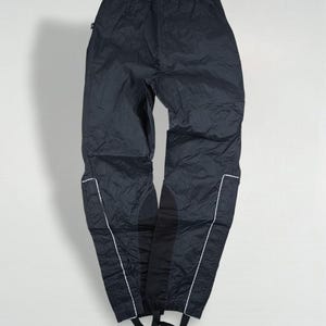 Könnte beinhalten: Schwarze Sporthose aus Nylon mit weißen Zierleisten entlang der Nähte. Die Hose hat Steigbügel an den Knöcheln und ist für Outdoor-Aktivitäten konzipiert. Das Material wirkt leicht und wasserabweisend.